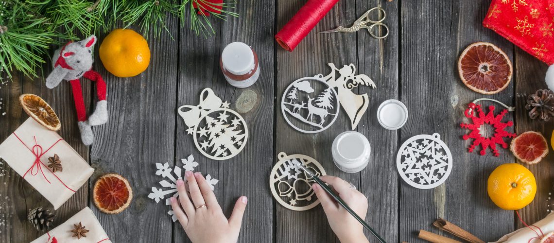 The girl makes wooden Christmas tree toys, draws white paint on a wooden table next to festive decorations.Christmas diy.Zero waste.Children's crafts. creative approach.leisure with children.flat lay