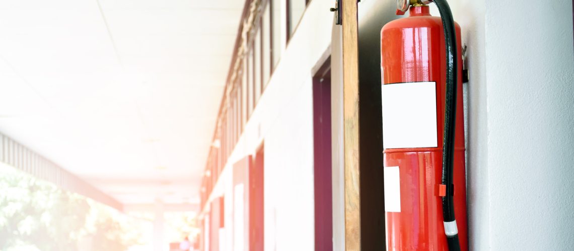 A red fire extinguisher is installed on a white cement wall in the front porch of the building to be used to extinguish a fire in the event of a building fire.