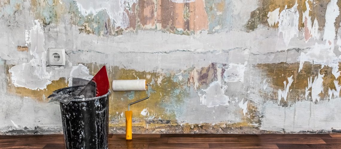 A bucket of construction tools and a paint roller sit on the floor against a shabby wall with putty stains and pieces of wallpaper in the room. Painting services and wallpapering in the apartment