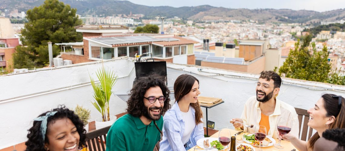 Young group of people enjoying dinner reunion at rooftop home. Millennial diverse friends social gathering reunited on terrace table, having fun celebrating barbecue party.