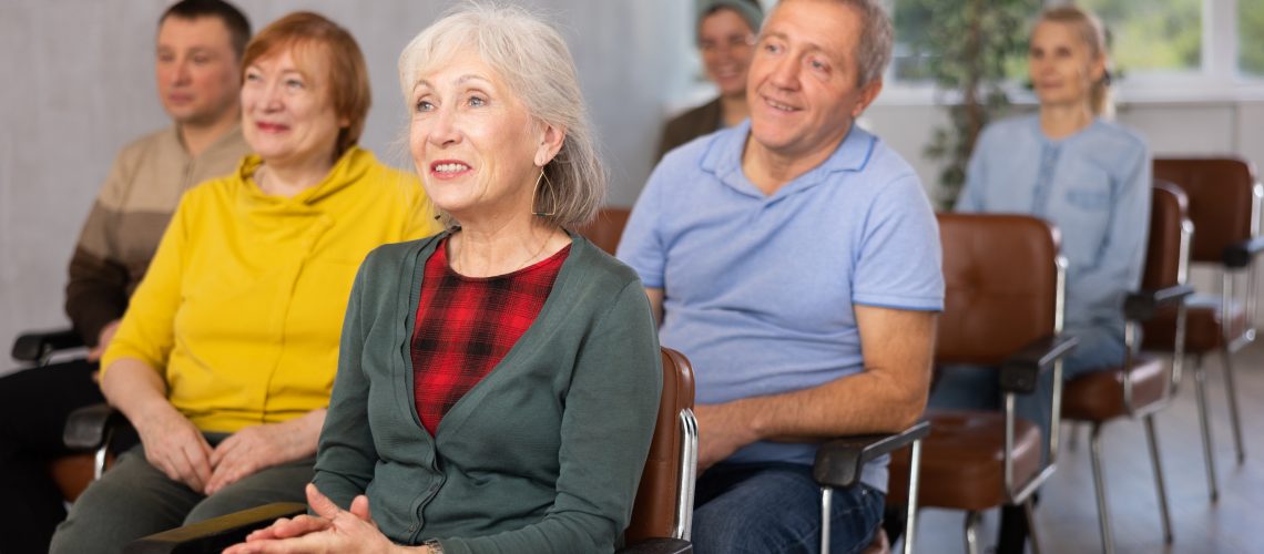 Interested aged woman sitting in auditorium with group, listening to lecture. Concept of learning program for seniors