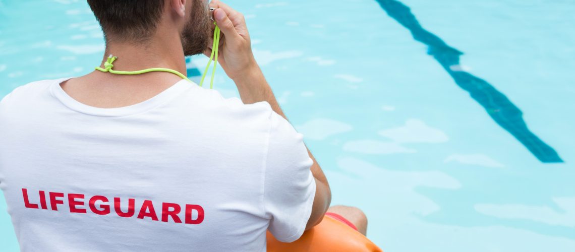 Rear view of lifeguard sitting on chair and blowing whistle at poolside